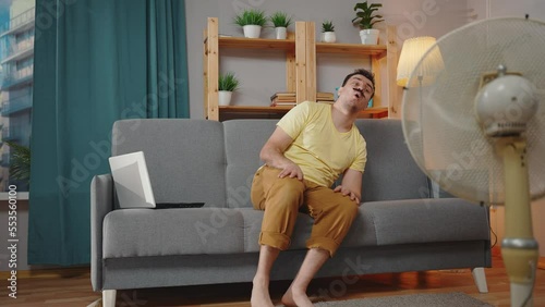 problem of hot weather , a male freak with a mustache suffering from heat and overheating in a hot summer is sitting on the sofa in the living room with a cooler fan