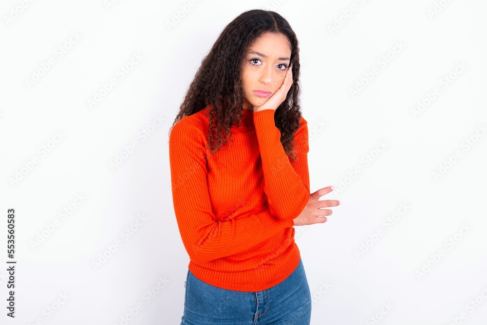 Very bored teen girl wearing knitted red sweater over white background holding hand on cheek while support it with another crossed hand, looking tired and sick. Very bored teen girl wearing knitted red sweater over white background holding hand on cheek while support it with another crossed hand, looking tired and sick.