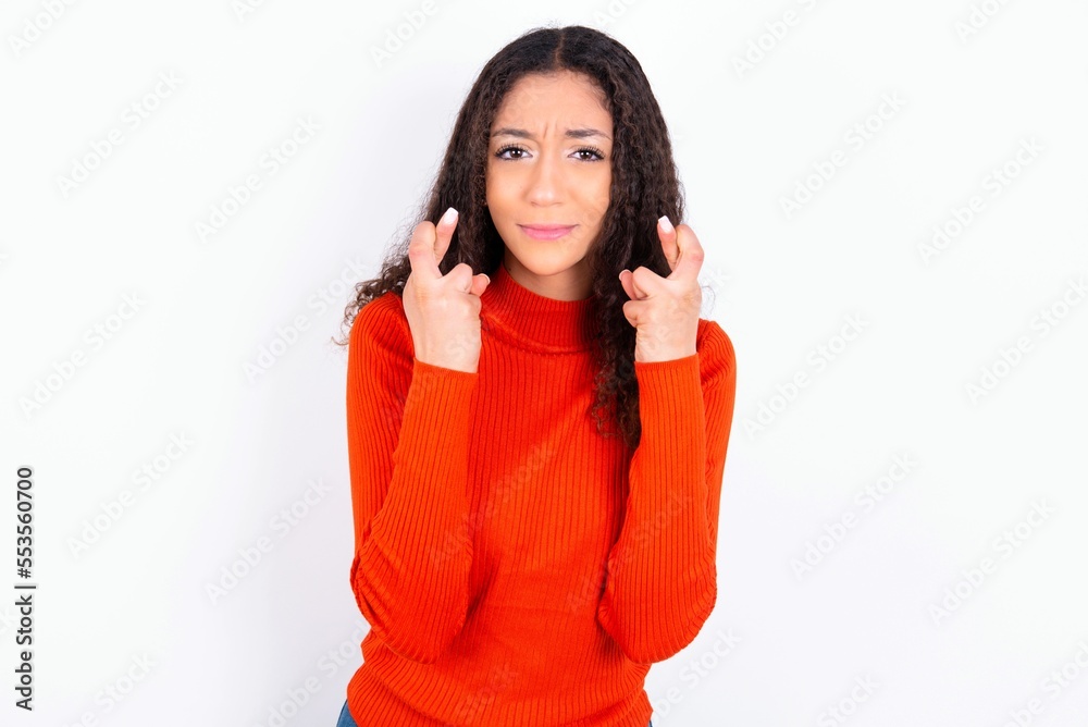 teen girl wearing knitted red sweater over white background holding fingers crossed with worried expression hoping boss didn't noticed mistakes at work. teen girl wearing knitted red sweater over white background holding fingers crossed with worried expression hoping boss didn't noticed mistakes at work.