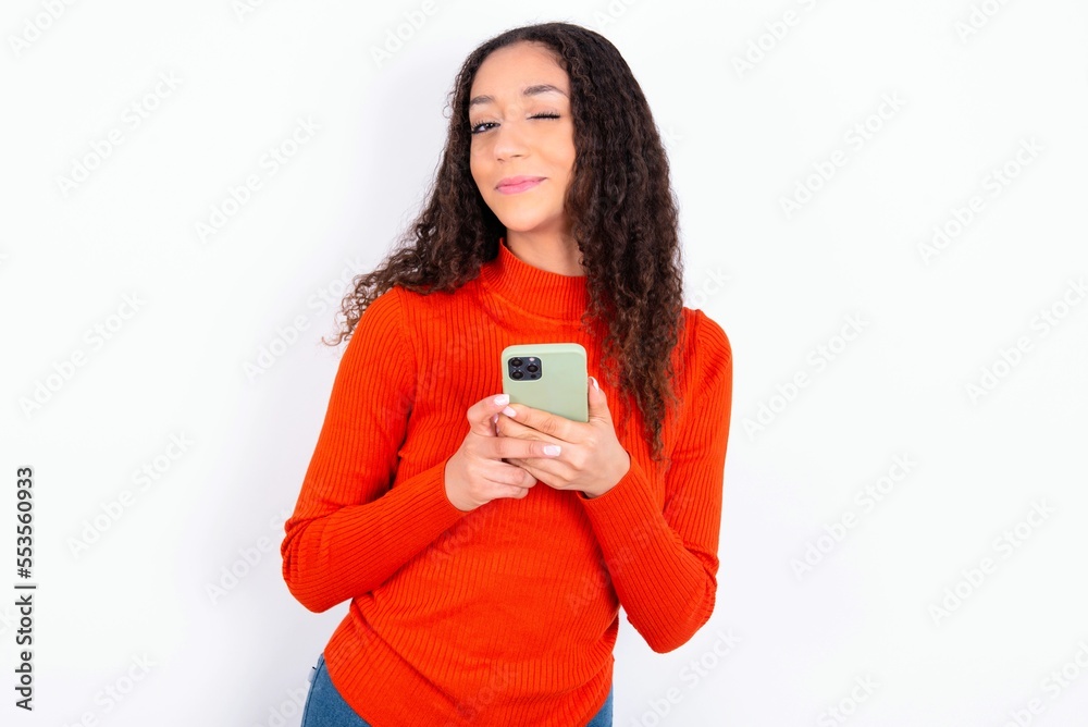 Pleased teen girl wearing knitted red sweater over white background using self phone and looking and winking at the camera. Flirt and coquettish concept. Pleased teen girl wearing knitted red sweater over white background using self phone and looking and winking at the camera. Flirt and coquettish concept.