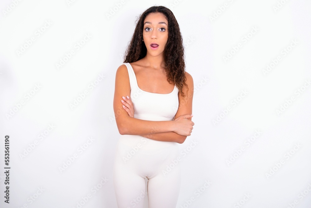 Shocked embarrassed teen girl with curly hair wearing white sport set over white background keeps mouth widely opened. Hears unbelievable novelty stares in stupor Shocked embarrassed teen girl with curly hair wearing white sport set over white background keeps mouth widely opened. Hears unbelievable novelty stares in stupor
