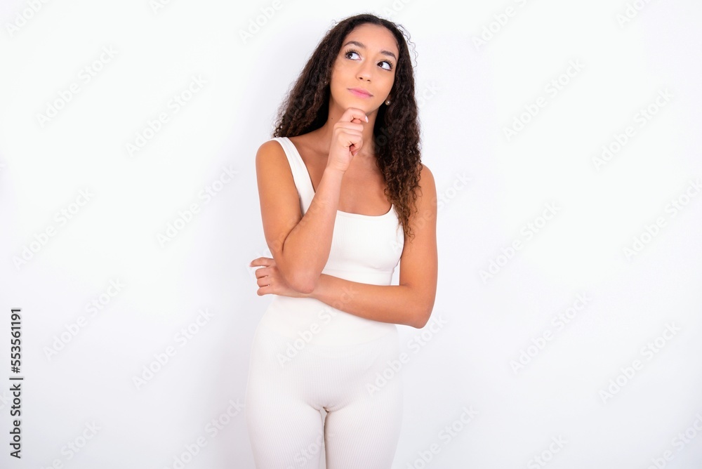 Thoughtful teen girl with curly hair wearing white sport set over white background holds chin and looks away pensively makes up great plan Thoughtful teen girl with curly hair wearing white sport set over white background holds chin and looks away pensively makes up great plan