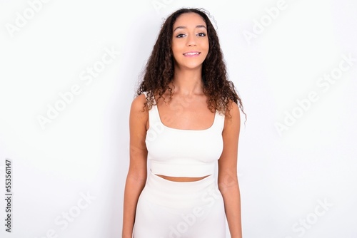 teen girl with curly hair wearing white sport set over white background with nice beaming smile pleased expression. Positive emotions concept