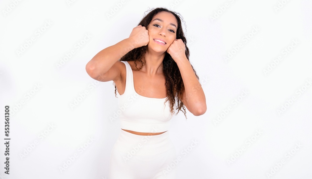 Happy teen girl with curly hair wearing white sport set over white background keeps fists on cheeks smiles broadly and has positive expression being in good mood Happy teen girl with curly hair wearing white sport set over white background keeps fists on cheeks smiles broadly and has positive expression being in good mood