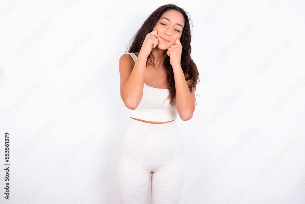 Pleased teen girl with curly hair wearing white sport set over white background with closed eyes keeps hands near cheeks and smiles tenderly imagines something very pleasant Pleased teen girl with curly hair wearing white sport set over white background with closed eyes keeps hands near cheeks and smiles tenderly imagines something very pleasant