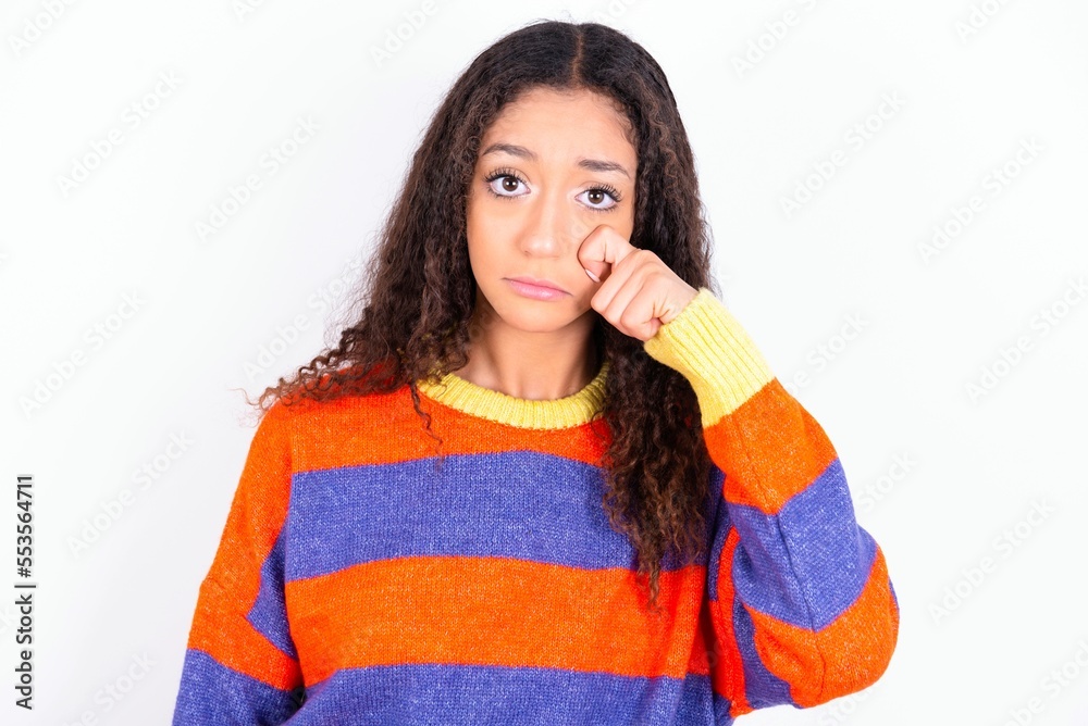 Disappointed dejected beautiful teen girl wearing knitted colourful sweater over white background wipes tears stands stressed with gloomy expression. Negative emotion Disappointed dejected beautiful teen girl wearing knitted colourful sweater over white background wipes tears stands stressed with gloomy expression. Negative emotion