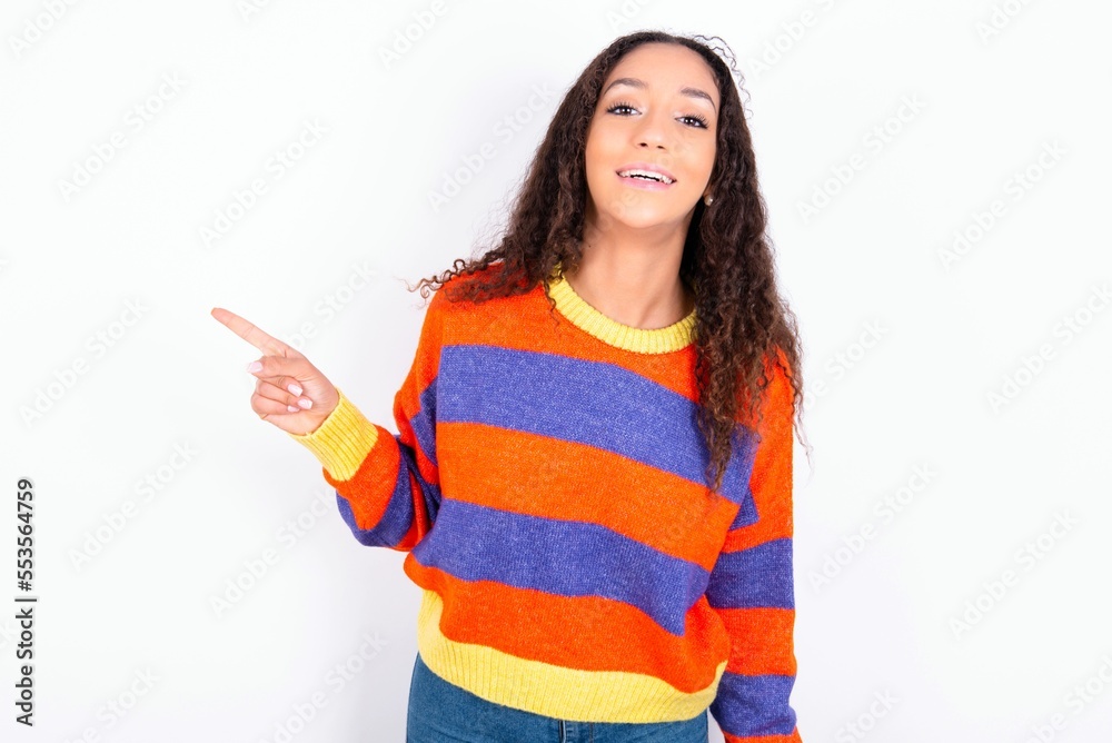 beautiful teen girl wearing knitted colourful sweater over white background laughs happily points away on blank space demonstrates shopping discount offer, excited by good news or unexpected sale. beautiful teen girl wearing knitted colourful sweater over white background laughs happily points away on blank space demonstrates shopping discount offer, excited by good news or unexpected sale.