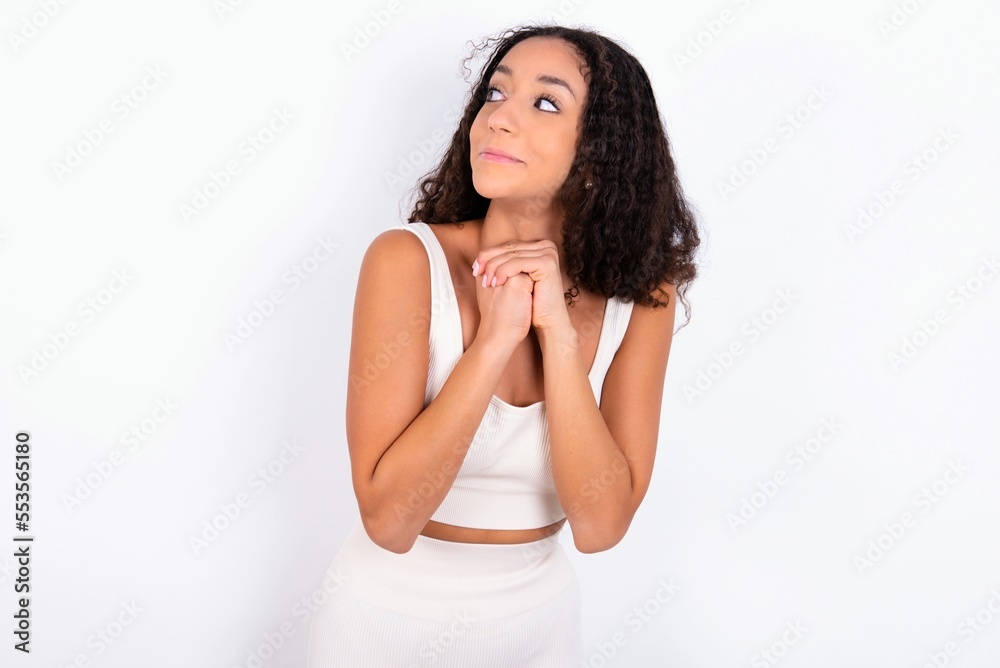 Curious teen girl with curly hair wearing white sport set over white background keeps hands under chin bites lips and looks with interest aside. Curious teen girl with curly hair wearing white sport set over white background keeps hands under chin bites lips and looks with interest aside.