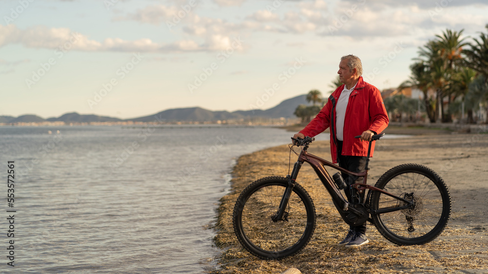 Fototapeta premium Senior man at the seaside with an electric mountain bike. sunny day at sunset. Copy space on the left. Concept of growing old without losing youth