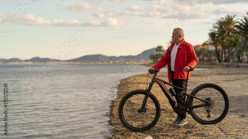 Senior man at the seaside with an electric mountain bike. sunny day at sunset. Copy space on the left. Concept of growing old without losing youth