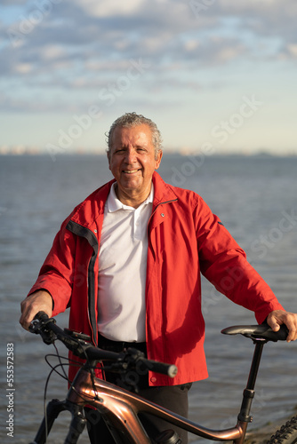 Senior man posing with his electric bike to the camera. Smiling man, behind is the sea. Portrait. Concept of growing old without losing youth
