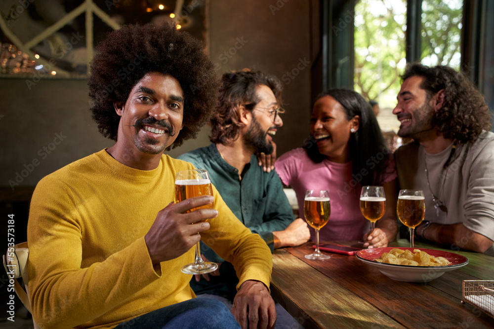 Copy space Portrait of smiling Afro hairstyle man looking at the camera and posing holding a beer while his cheerful friends laugh and have fun at the bar in the background. People posing to camera.