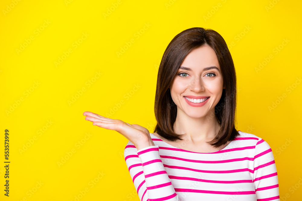Photo portrait of stunning young lady smiling hold palm copyspace promotion wear trendy striped garment isolated on yellow color background Photo portrait of stunning young lady smiling hold palm copyspace promotion wear trendy striped garment isolated on yellow color background