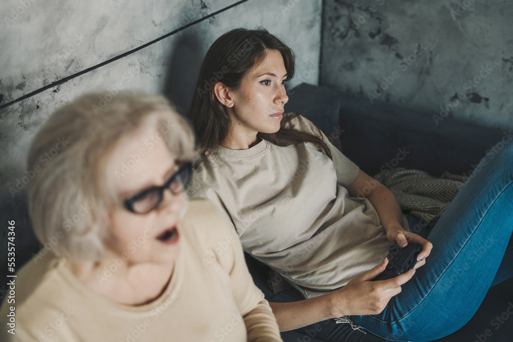 Cheerful adult woman playing video games with her senior mother while sitting on the sofa. Happy lifestyle. People lifestyle. Family home leisure.