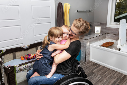 A mother in a wheelchair holds her baby and young daughter doing a group hug in a disability equipped bathroom; Edmonton, Alberta, Canada photo