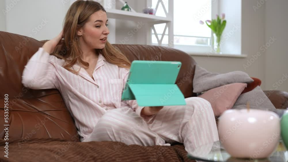 Beautiful woman with toothy smile messaging online using video chat on digital tablet sitting on sofa in living room. Portrait of positive charming Caucasian lady in pajama enjoying weekend morning Beautiful woman with toothy smile messaging online using video chat on digital tablet sitting on sofa in living room. Portrait of positive charming Caucasian lady in pajama enjoying weekend morning