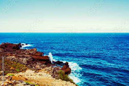 Scenic view of the ocean and the Nakalele Blowhole against a blue sky with the rugged, rocky coastline in the foreground; Maui, Hawaii, United States of America photo