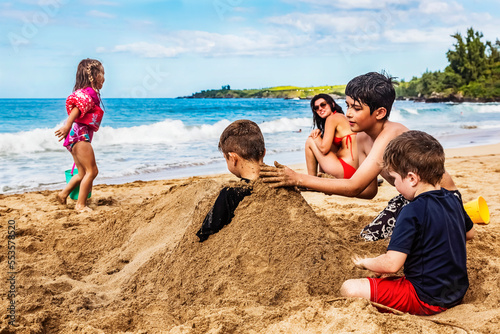 Mother sitting on D. T. Fleming Beach at the water's edge and watching her children playing and burying a sibling in the sand; Kapalua, Maui, Hawaii, United States of America photo