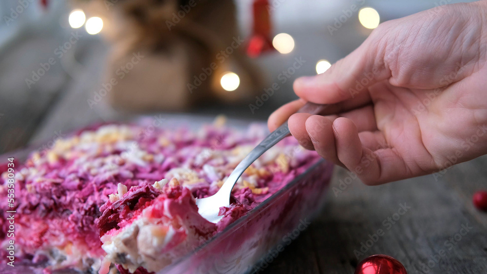 Homemade New Year is a herring salad under a fur coat on a wooden background. A man's hand lays a Russian traditional Christmas salad with a spoon.