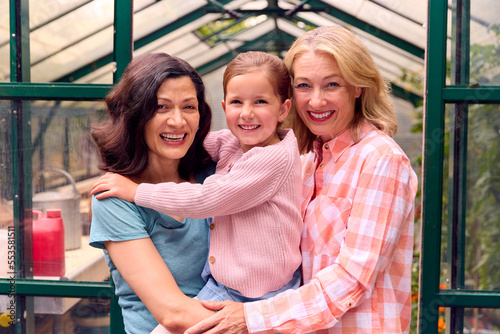 Portrait Of Same Sex Family With Two Mature Mums Hugging Daughter As They Garden In Greenhouse