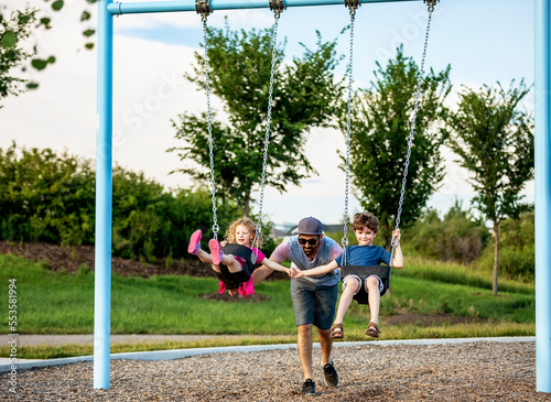 A father pushes a young girl and boy on swings on a swing set at a playground; St. Albert, Alberta, Canada
