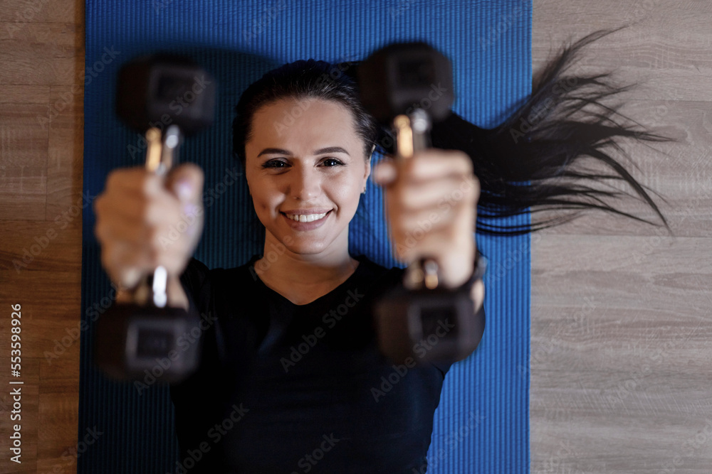 Top view portrait brunette sports woman lying on floor mat holding dumbbells exercising at gym Top view portrait brunette sports woman lying on floor mat holding dumbbells exercising at gym