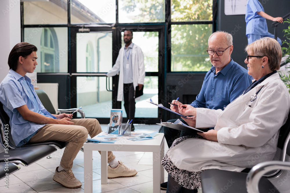 Senior doctor standing beside elderly patient discussing health care treatment during checkup visit appointment in hospital waiting area. Old man filling insurance report before start consultation