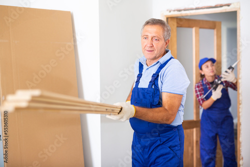 Caucasian middle-aged man builder in overall carrying wooden planks while working in construction site inside apartment. © JackF