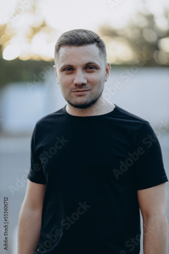 Young man in black t-shirt smiling happy standing at the city outdoors on a sunny evening. Handsome young man face - close up