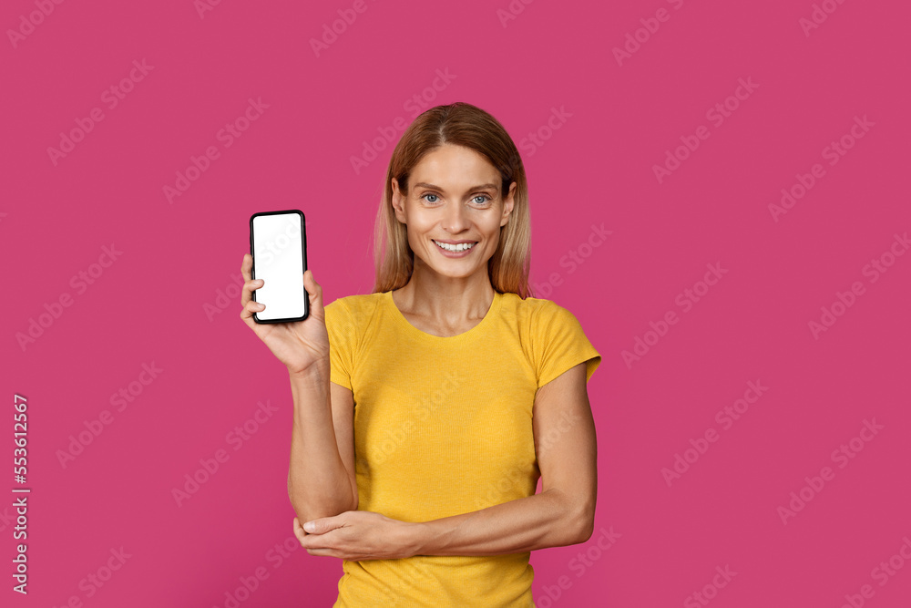 Cheerful confident mature caucasian blonde female in yellow t-shirt shows smartphone with blank screen Cheerful confident mature caucasian blonde female in yellow t-shirt shows smartphone with blank screen