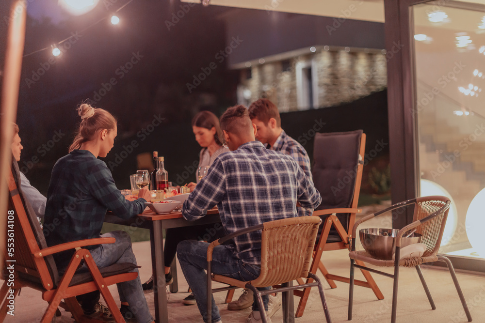 Fototapeta premium A group of young diverse people having dinner on the terrace of a modern house in the evening. Fun for friends and family. Celebration of holidays, weddings with barbecue.