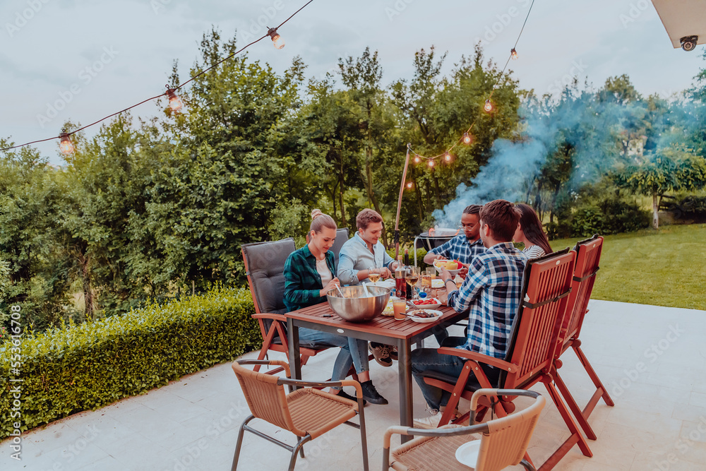 © .shock - A group of young diverse people having dinner on the terrace of a modern house in the evening. Fun for friends and family. Celebration of holidays, weddings with barbecue.