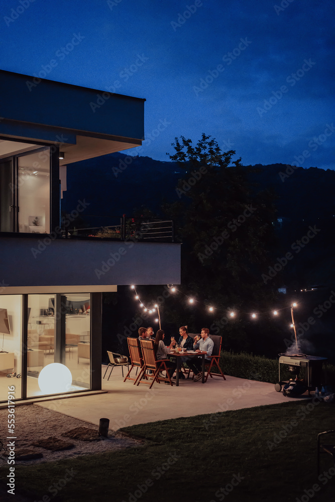 © .shock - A group of young diverse people having dinner on the terrace of a modern house in the evening. Fun for friends and family. Celebration of holidays, weddings with barbecue.