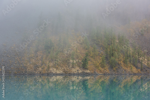 Simple meditative landscape with forest hill reflection in tranquil turquoise lake in fog. Mossy shore with firs in fading autumn colors reflected in mirror mountain lake. Autumn shore of alpine lake.