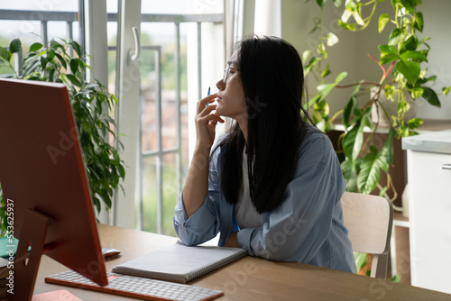 Procrastination in online learning. Dreamy Asian student girl getting distracted while studying remotely at home, sitting at desk with computer looking out window, having no motivation to study