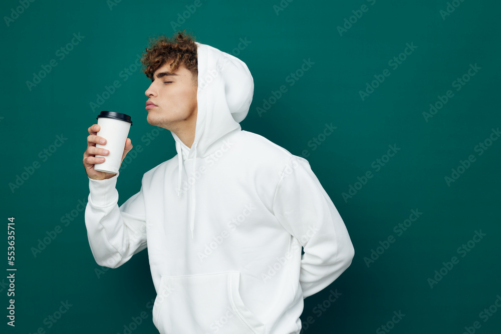 a handsome man stands on a green background in a white hoodie holding a glass in his hand and reaches out to him with his face inhaling the aroma of the drink. Horizontal Studio Photography