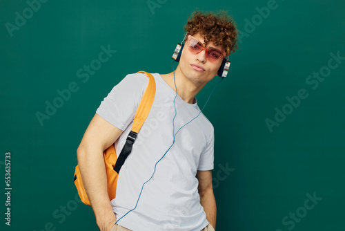 a joyful man, a teenager, stands on a green background listening to music in headphones with pink glasses on his face and looks at the camera