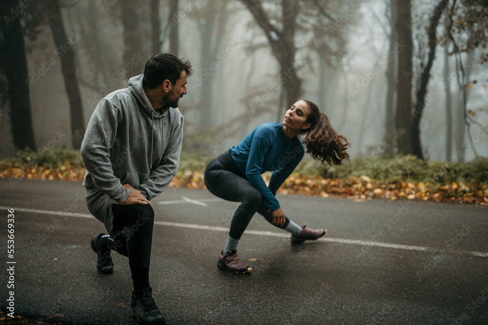 Dedicated and cheerful fit woman and man having outdoor jogging and workout in the foggy forest. Man and woman looking at each other and warming up legs before jogging Dedicated and cheerful fit woman and man having outdoor jogging and workout in the foggy forest. Man and woman looking at each other and warming up legs before jogging
