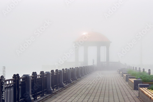 Architectural landscape of the rotunda on the embankment in the fog. Sun glare on the transparent dome of the gazebo above the blurred silhouette of a man. Fence and benches with flower beds photo