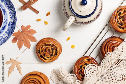 Buns with cinnamon and raisins cool on the grill under a lace napkin, on a light background. With a teapot, autumn leaves and a cinnamon stick photo