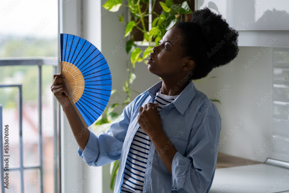 Young casual African American woman fanning herself with hand fan experiencing health problems due to hot weather. Black girl in blue shirt lookina at window standing in room with home plants Stock Fo...