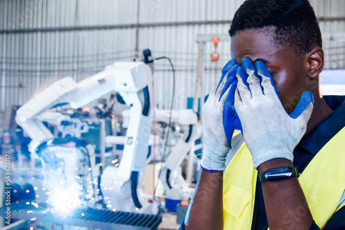 African american male worker staring at glare during steel welding, eye pain irritation tearful eyes hands covering face preventing flares sparks from electric welding with robotic arm in factory.