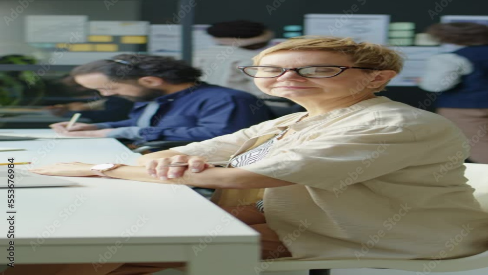 Vertical shot of short haired businesswoman in eyeglasses sitting at desk and posing for camera while working in office Vertical shot of short haired businesswoman in eyeglasses sitting at desk and posing for camera while working in office