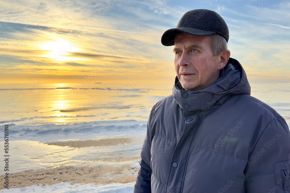 Portrait of beautiful happy elderly senior man, retired person walking at winter cold beach at nature, at cold frosty weather at sunset in scarf and hat, smiling