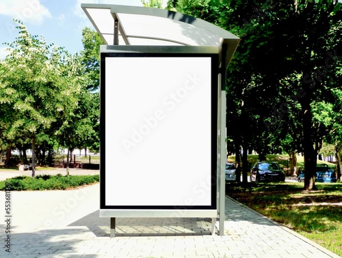 bus shelter at a busstop. blank billboard ad display. empty white lightbox sign. urban setting. old European street. city transit station. glass and aluminum frame structure. outdoor. green trees.