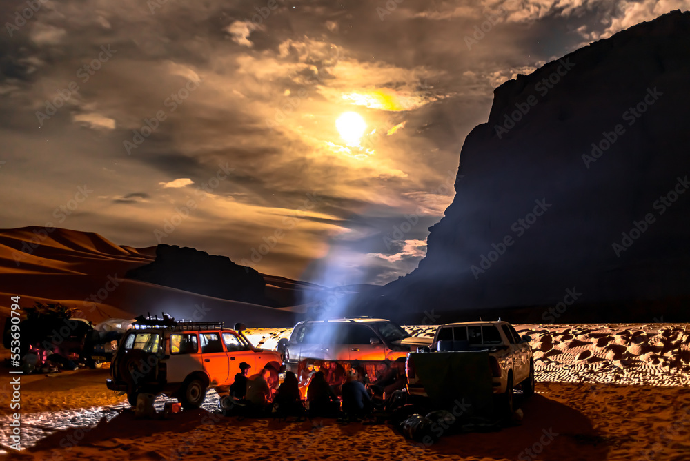 Campfire group and cars in Sahara Desert by night. 4X4 vehicules parked, Unrecognizable tourists and local berber people sitting around a camp-fire in Tadrart Rouge. Cloudy sky and full moon moonlight