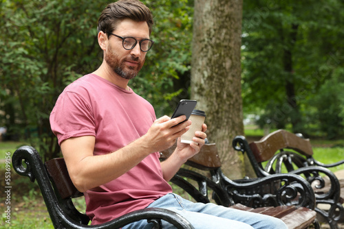 Handsome man with coffee using smartphone in park, space for text