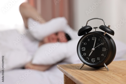 Man covering his ears with pillows in bedroom, focus on alarm clock. Space for text