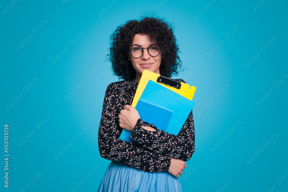 Front view portrait of cheerful young brunette business woman wearing eyeglasses isolated on blue background studio. Hold notebooks, looking at camera. Front view portrait of cheerful young brunette business woman wearing eyeglasses isolated on blue background studio. Hold notebooks, looking at camera.