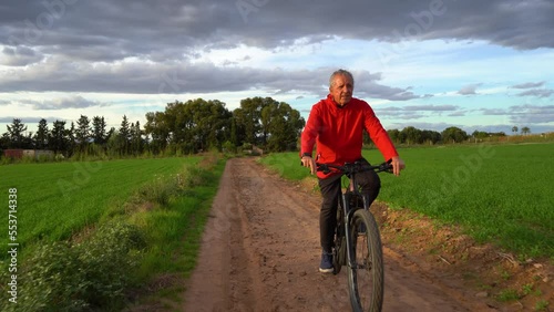 Senior man on electric mountain bike doing sports through a green field. He is on a road and in the background a wooded area and clouds. Cloudy day at sunset. Concept seniors in active, enjoying life
